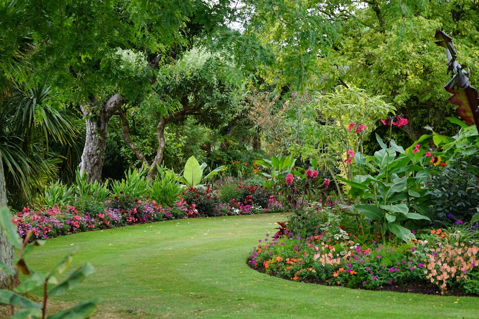 A lush garden scene featuring a well-maintained, vibrant green lawn bordered by a variety of flowering plants and shrubs. On the right side, tall pink lilies with broad green leaves stand amidst clust