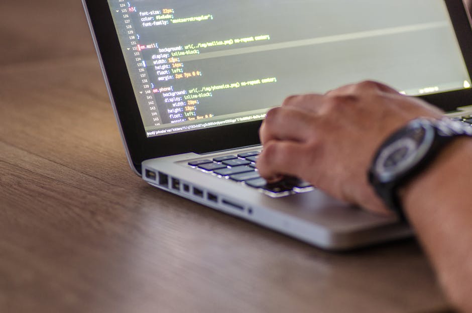 A close-up view of a person's hand typing on a silver laptop keyboard placed on a wooden desk. The screen displays lines of colorful code or programming language, with a dark background and brightly c
